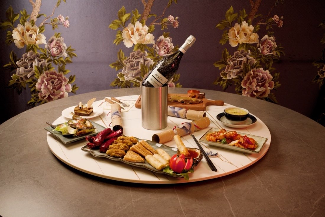 A banquet spread on a lazy susan with prosecco, against embroidered floral wallpaper