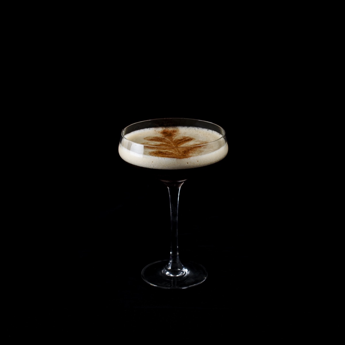 Espresso martini in a coupe glass on pure black, dusted with a leaf of cocoa