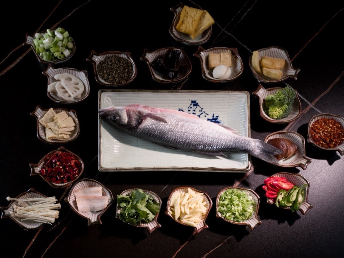 Whole sea bass on a painted platter ringed by small dishes of hotpot ingredients on dark stone