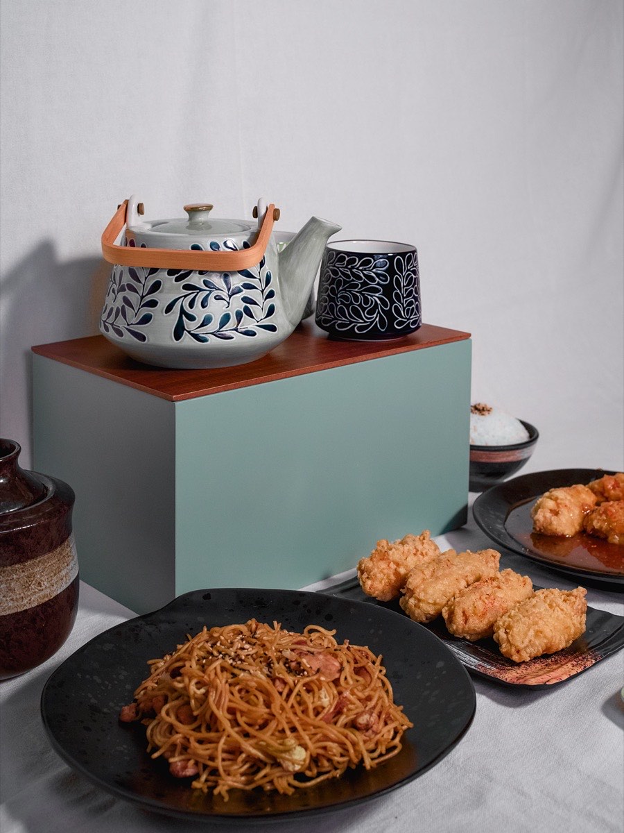 Patterned teapot and cup on a colour-blocked plinth beside noodles and tempura